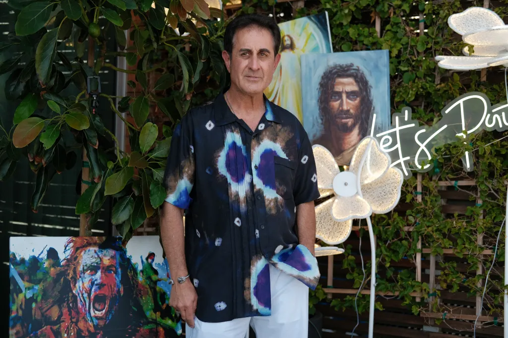 Bruce Lion, wearing a tie-dye shirt and white pants, stands before two paintings and a neon 