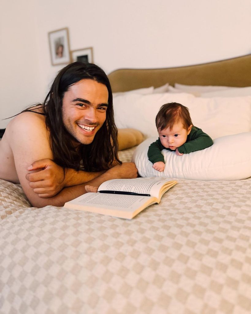 Derek Richard Thomas lying shirtless on a bed with an open book, smiling, next to his baby Lou.
