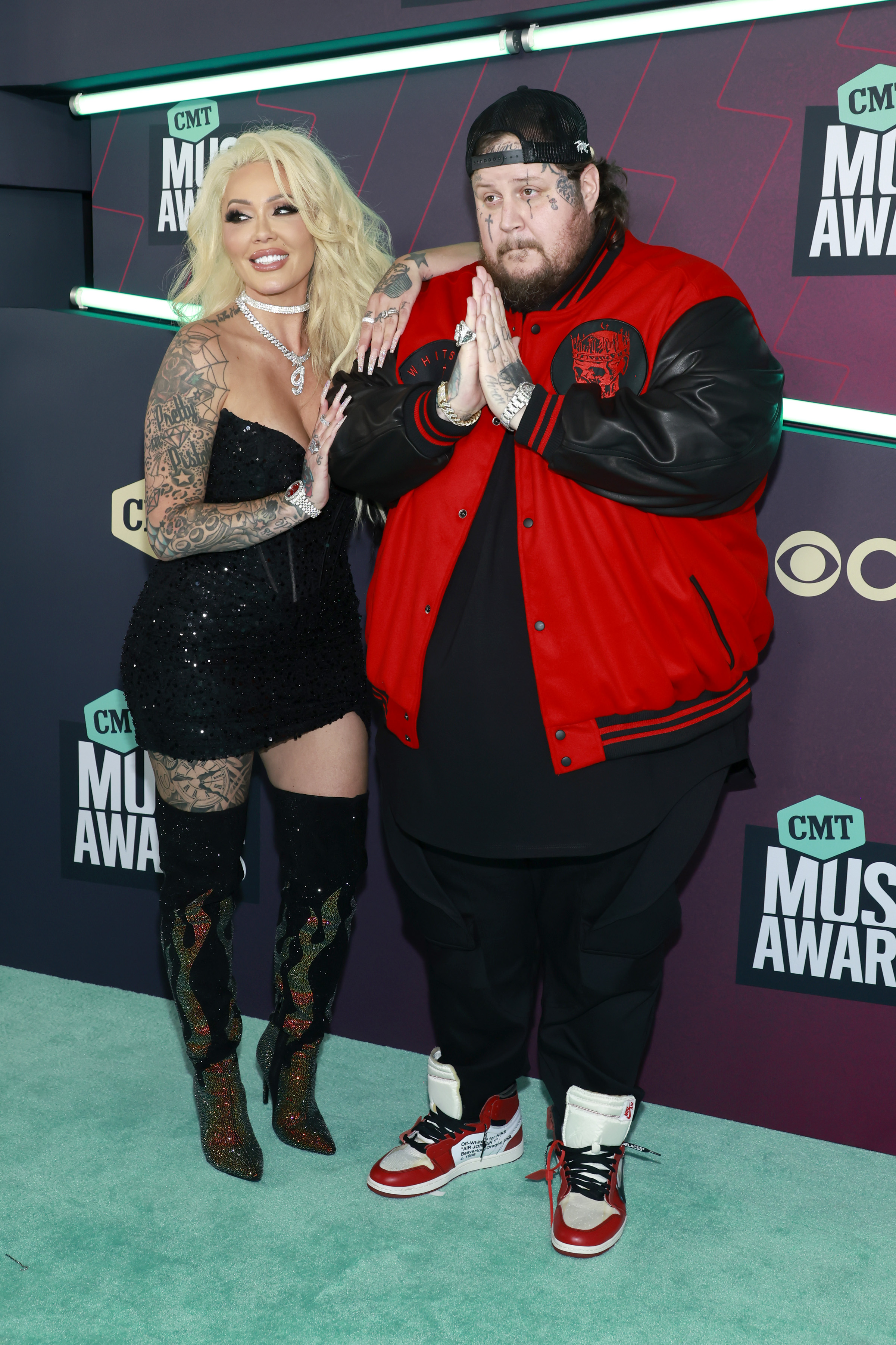 Bunnie Xo, in a sparkling black dress and thigh-high boots, and Jelly Roll, in a red and black varsity jacket, pose on the red carpet.