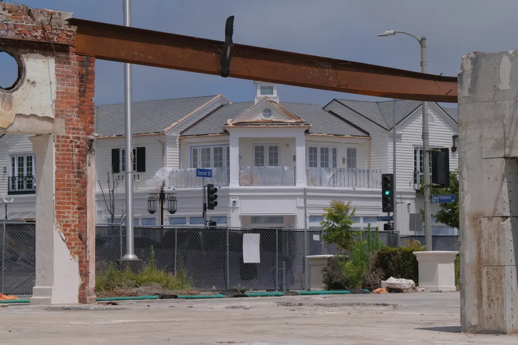 A white building with a balcony behind a fence, with damaged brick and concrete structures in the foreground.