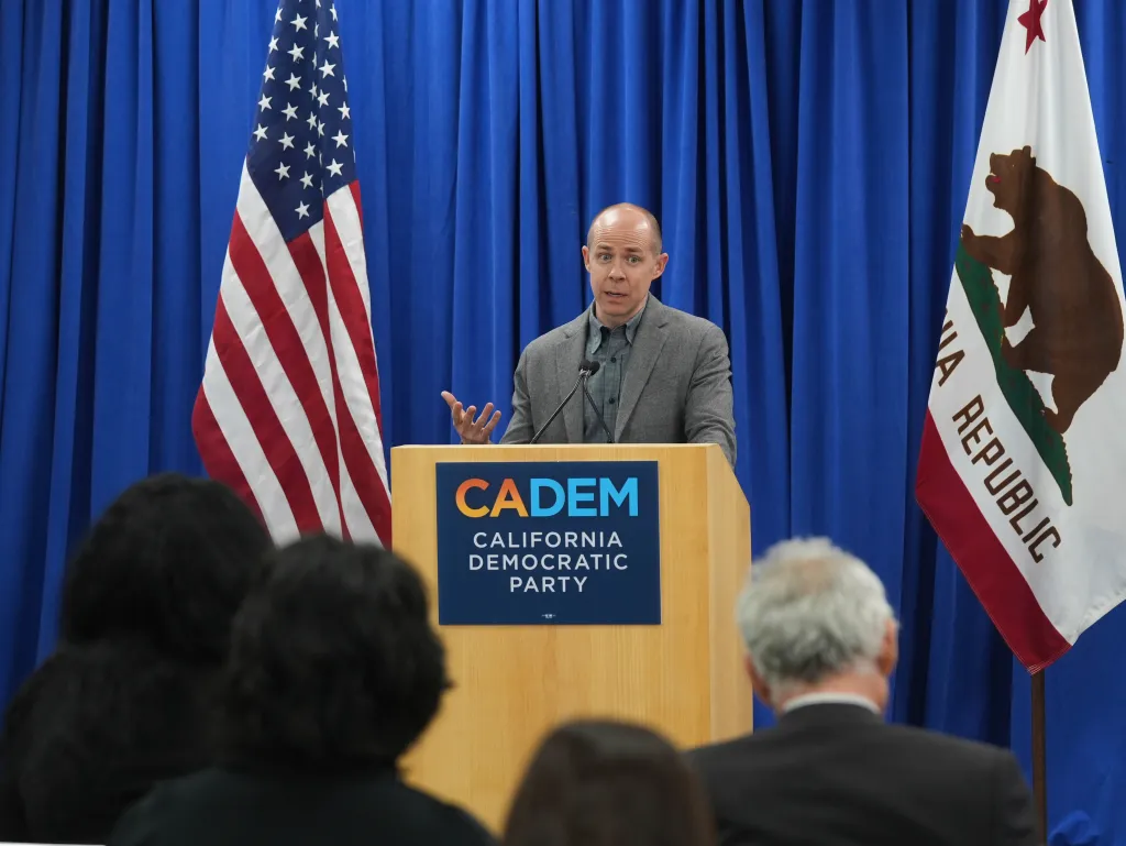 Rusty Hicks speaks at a podium with American and California state flags behind him.