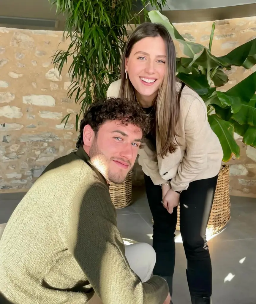 A man and woman pose for a photo in front of a stone wall and plants.