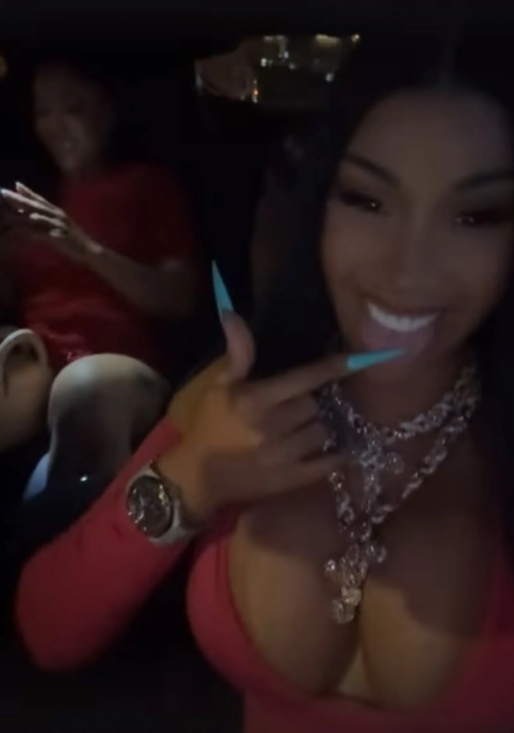 Cardi B in a red dress smiling and sticking her tongue out, with a second person visible behind her in the dark.