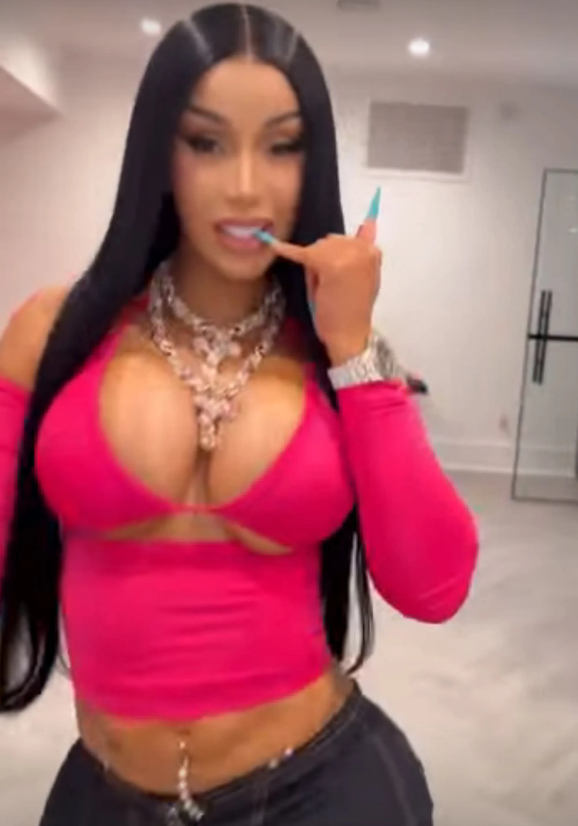 Cardi B in a racy pink top, revealing her cleavage and midriff, with long black hair, a chunky diamond necklace, and long blue fingernails.