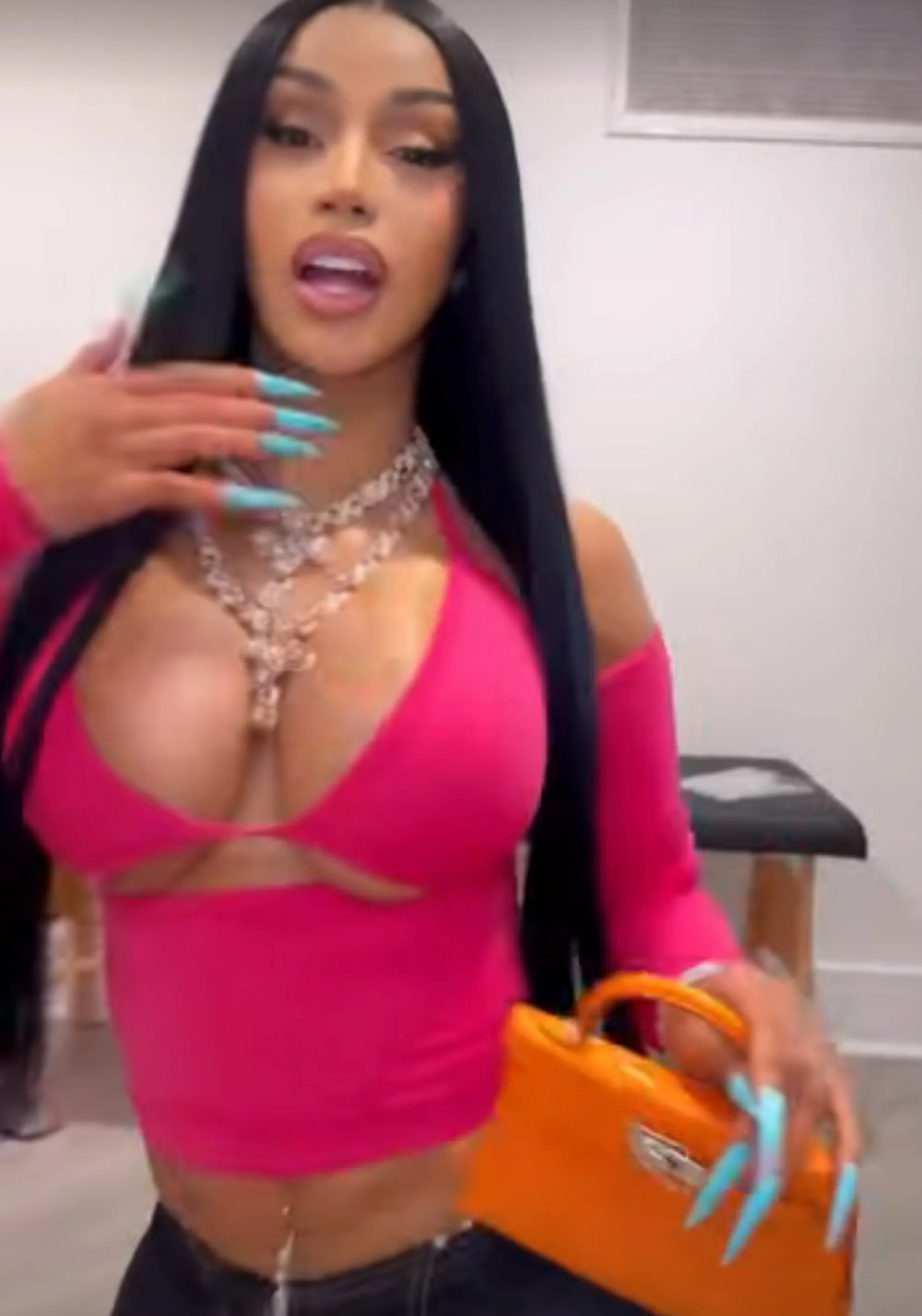 Cardi B in a pink top, holding an orange purse, with her long dark hair falling over her shoulders.