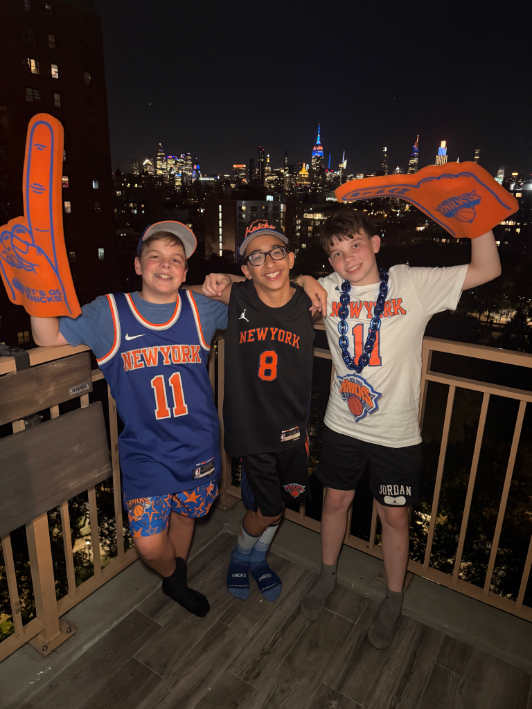 Three young boys on a balcony in New York City at night, wearing Knicks jerseys and holding foam fingers.
