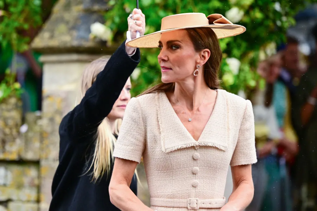 Catherine, Princess of Wales, wearing a tweed dress and a straw hat, looks right while an umbrella is held over her.