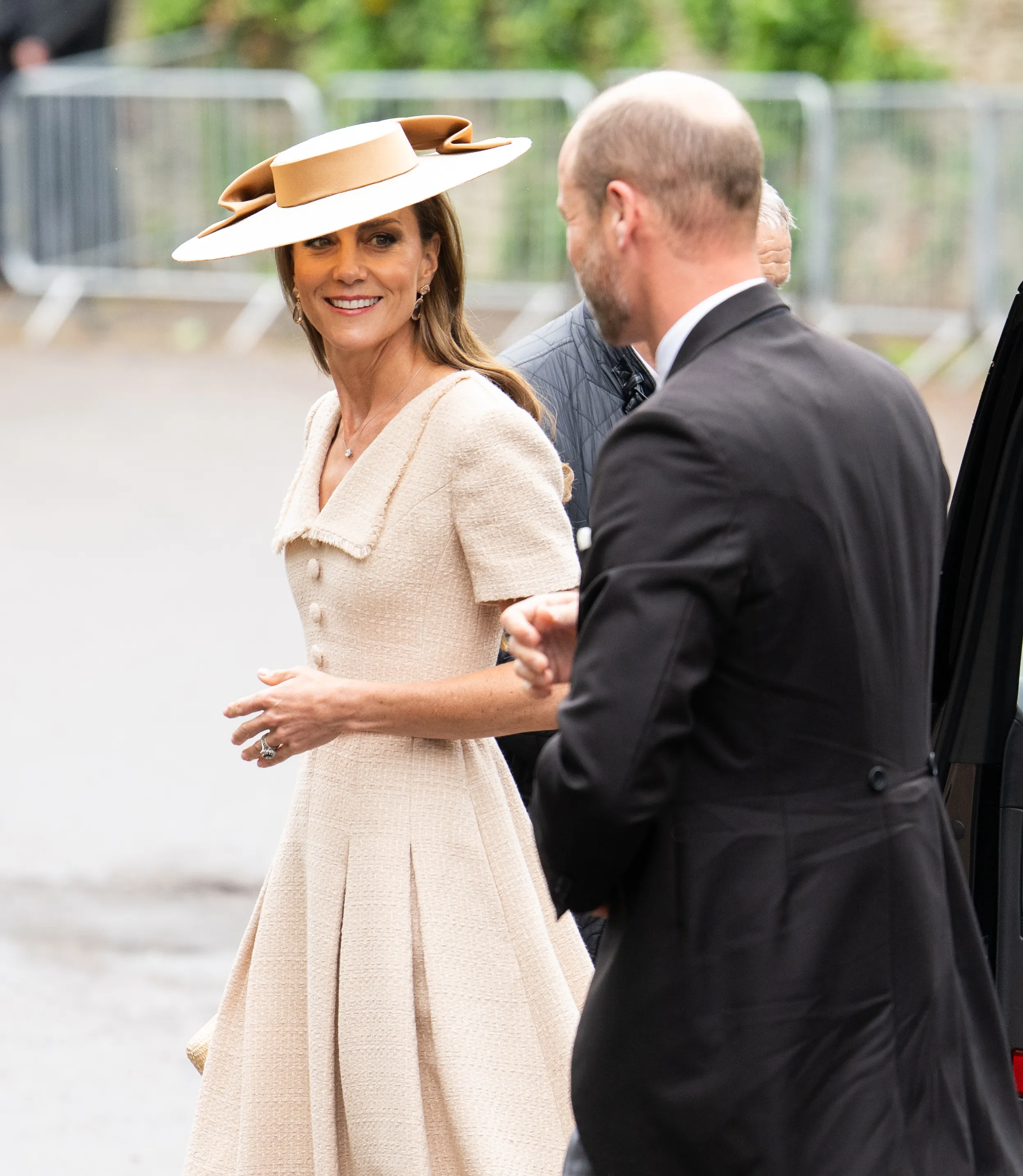 Kate Middleton in a cream dress and a matching wide-brimmed hat, smiling as she is assisted out of a car at a wedding.