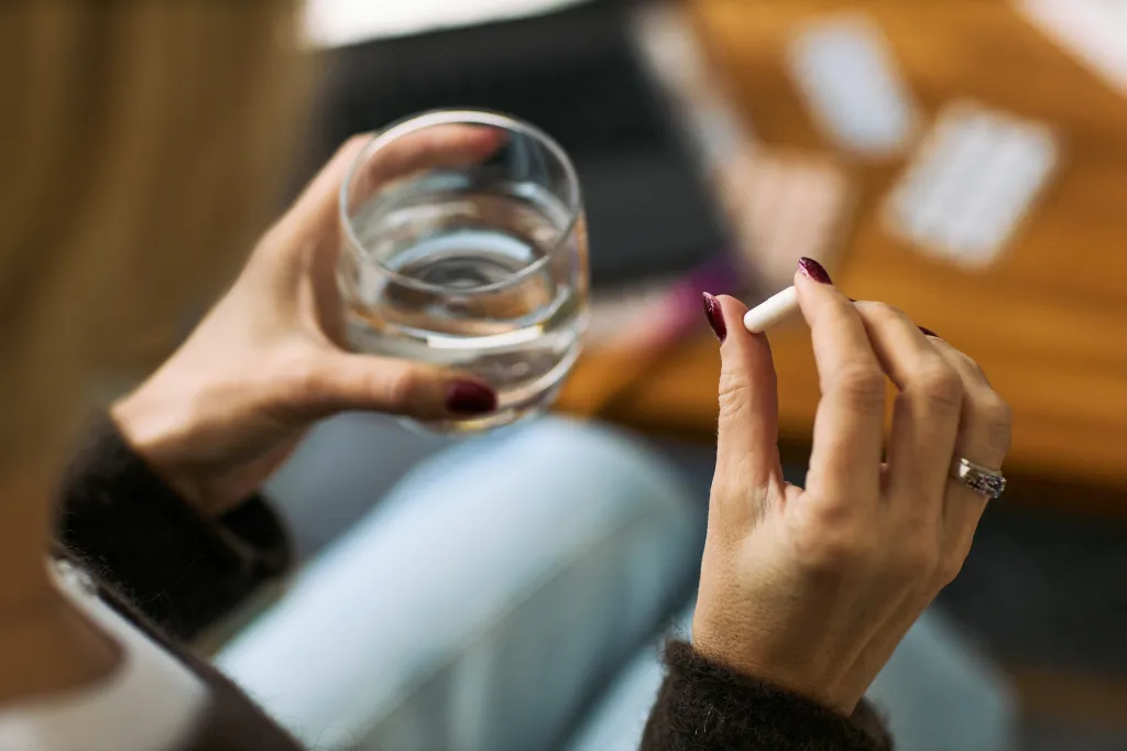 A woman's hands, with burgundy nail polish, hold a glass of water and a white pill.