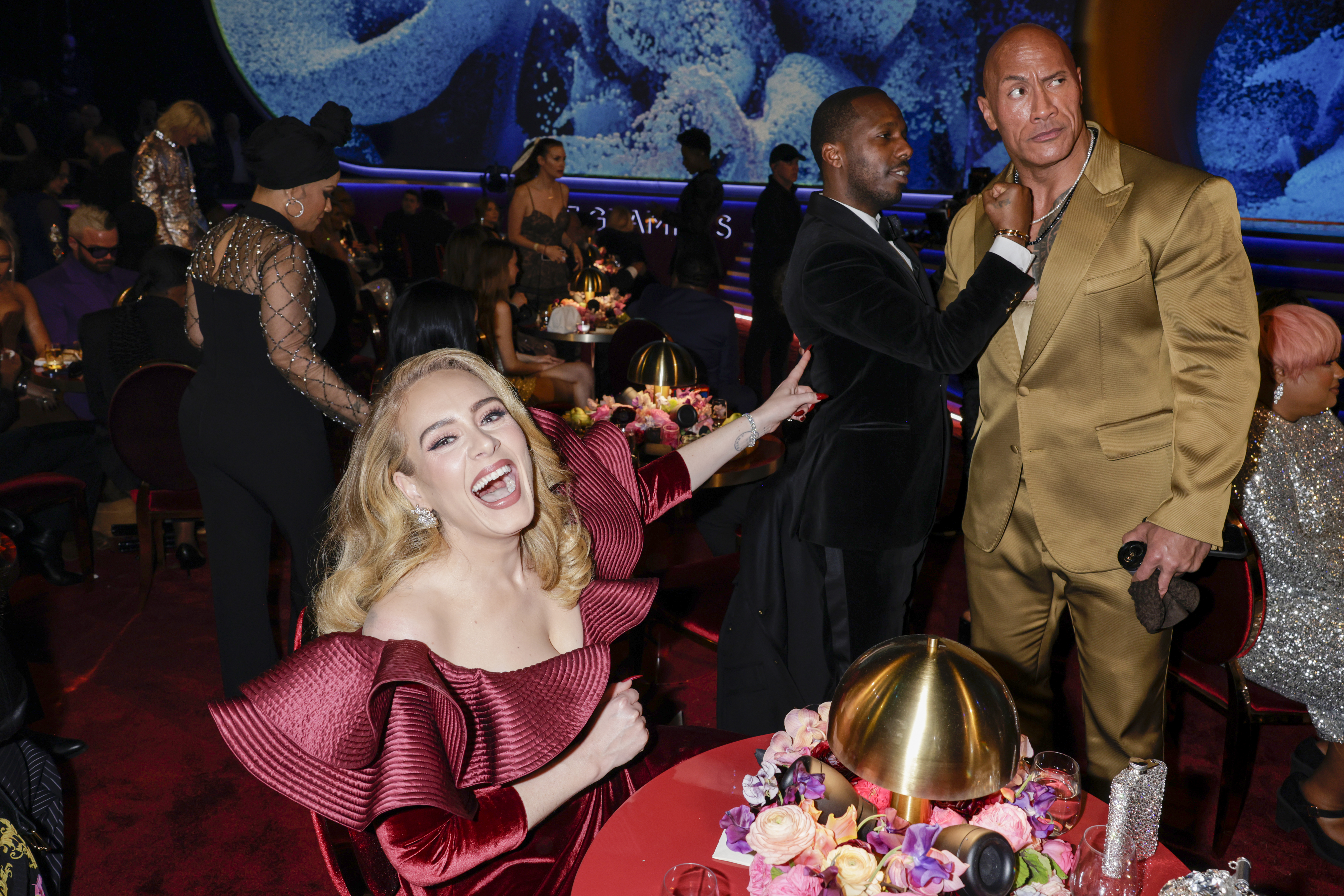 Adele laughs as she points at Dwayne Johnson, who stands beside Rich Paul at the Grammy Awards.
