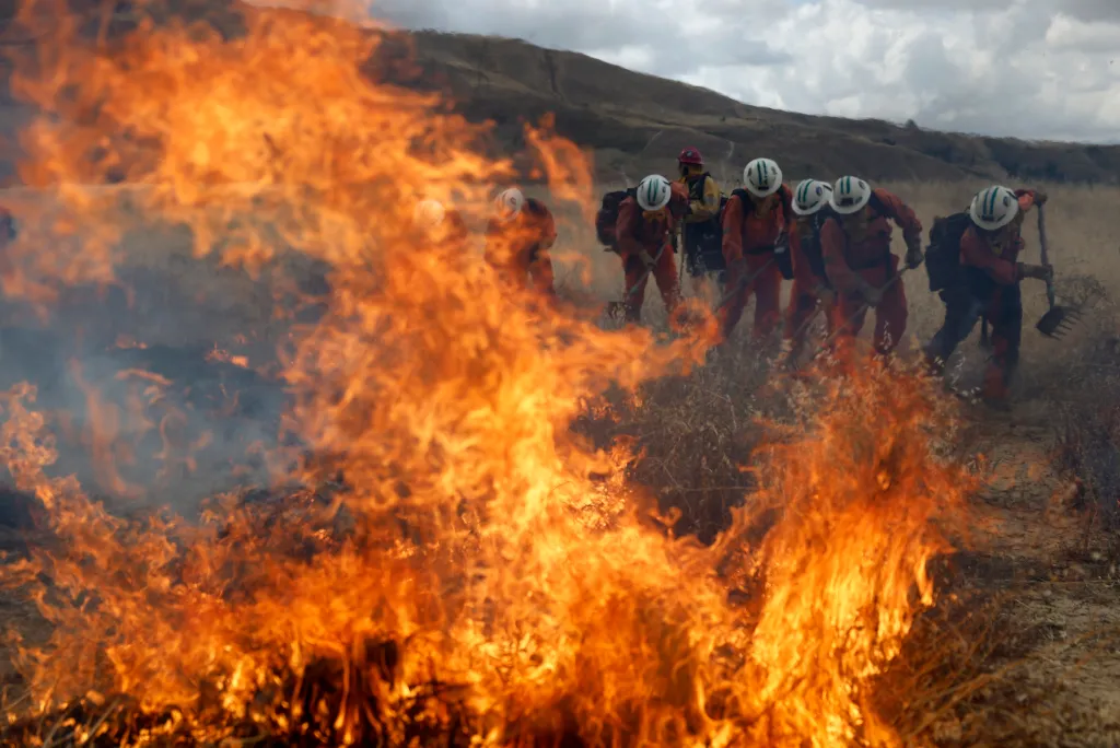 Inmates in protective gear fight a wildfire with tools, a hill and cloudy sky in the background.
