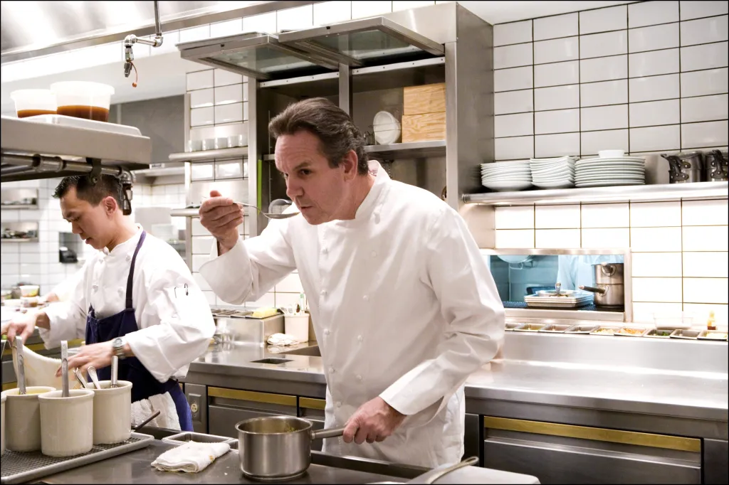 Chef Thomas Keller tasting food from a pot in a professional kitchen while another chef prepares food in the background.