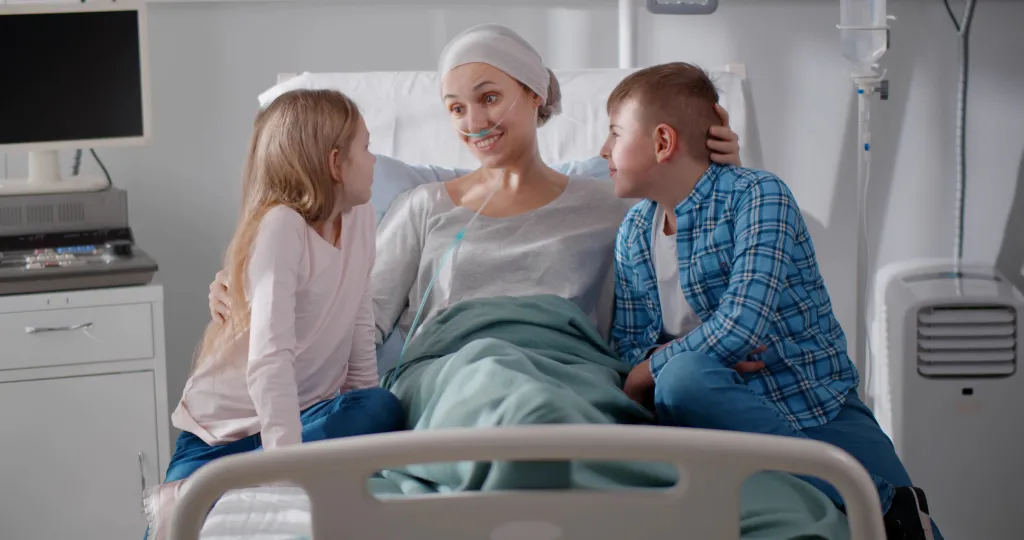 A mother in a hospital bed with a headscarf and nasal cannula smiles at her two children who are visiting her.