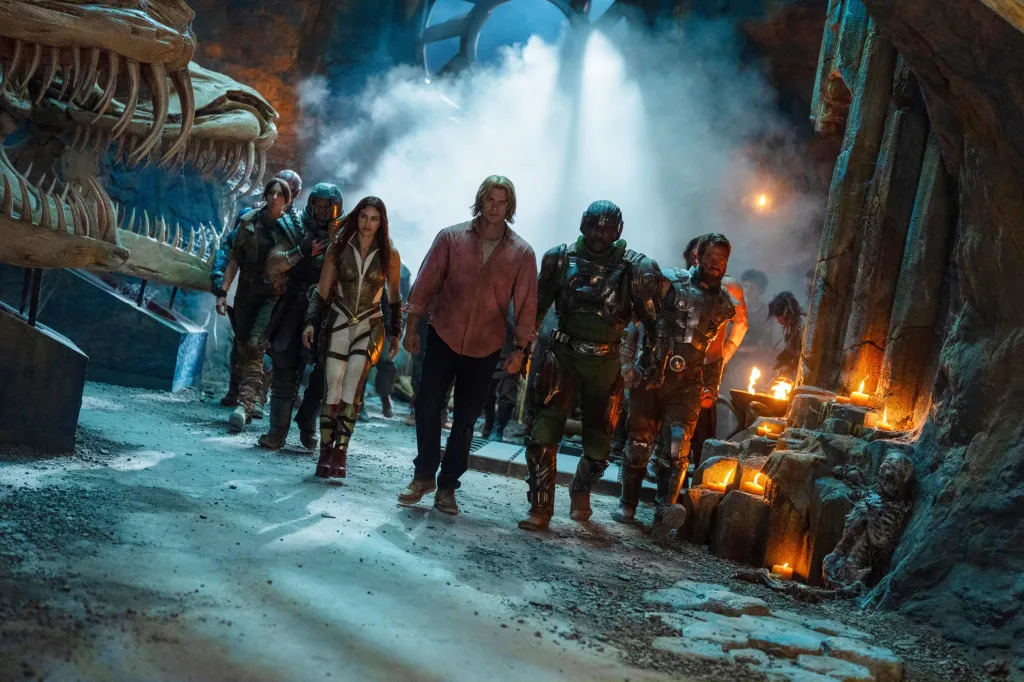 Cast members of Masters of the Universe walking through a cavern with a large skeletal jaw to the left, smoke filling the air, and lit candles to the right.