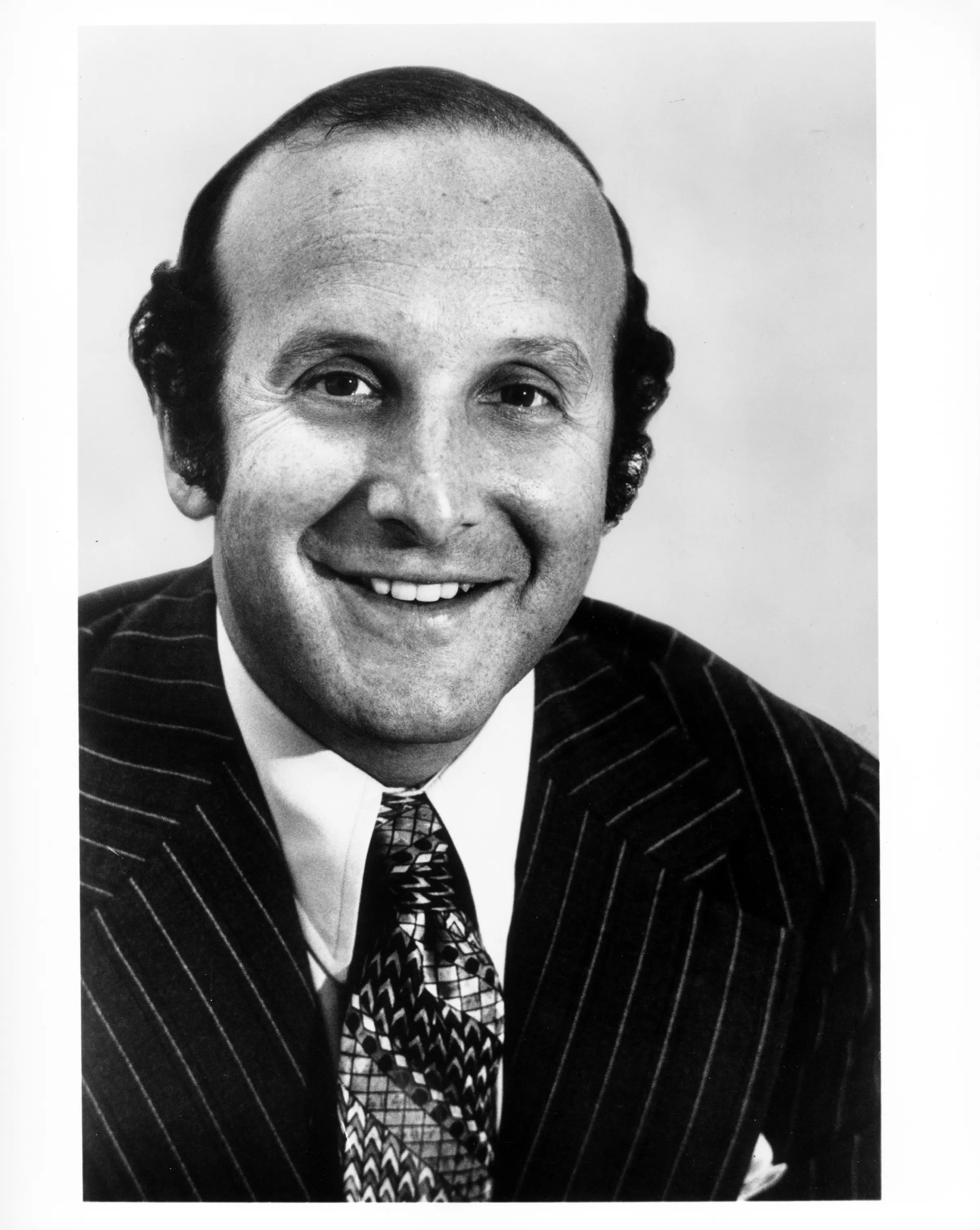 Black-and-white portrait of Clive Davis smiling at the viewer, wearing a pinstriped suit jacket and patterned tie.