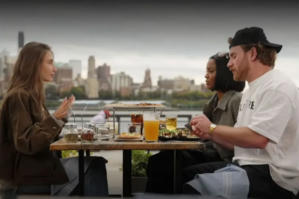 Amanda Batula, Ciara Miller, and West Wilson having a meal outdoors with a city skyline in the background.