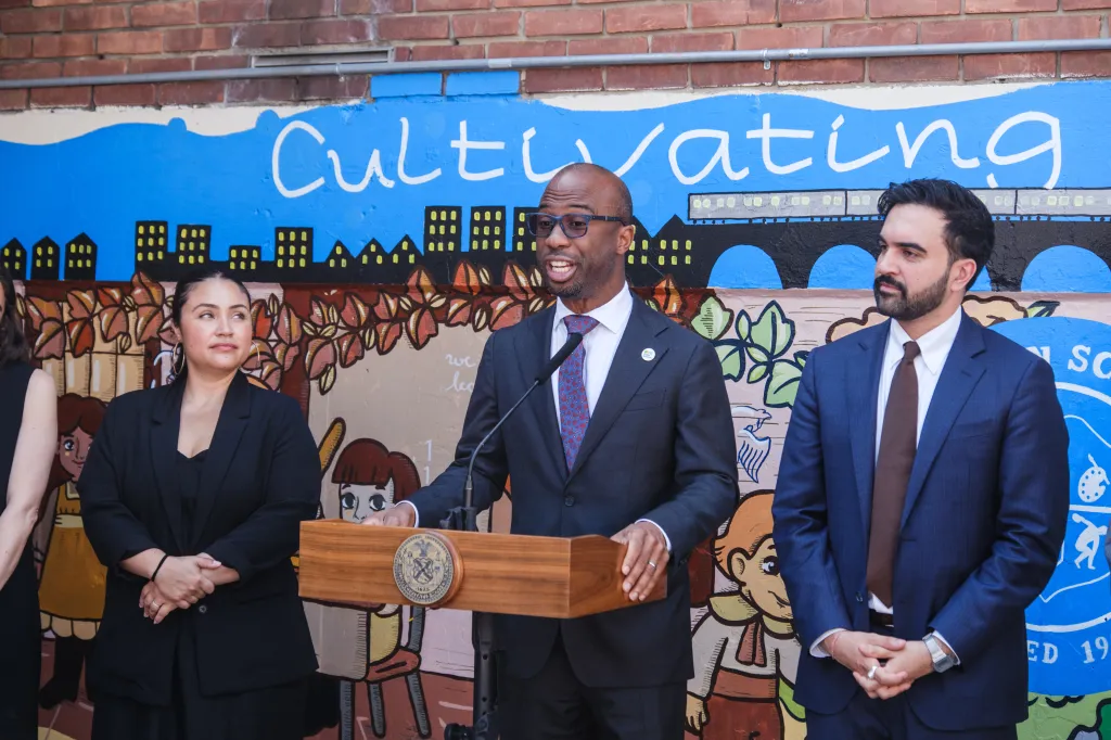 DOE Chancellor Kamar Samuels speaking at a podium alongside City Councilwoman Jennifer Gutierrez and NYC Mayor Zohran Mamdani.