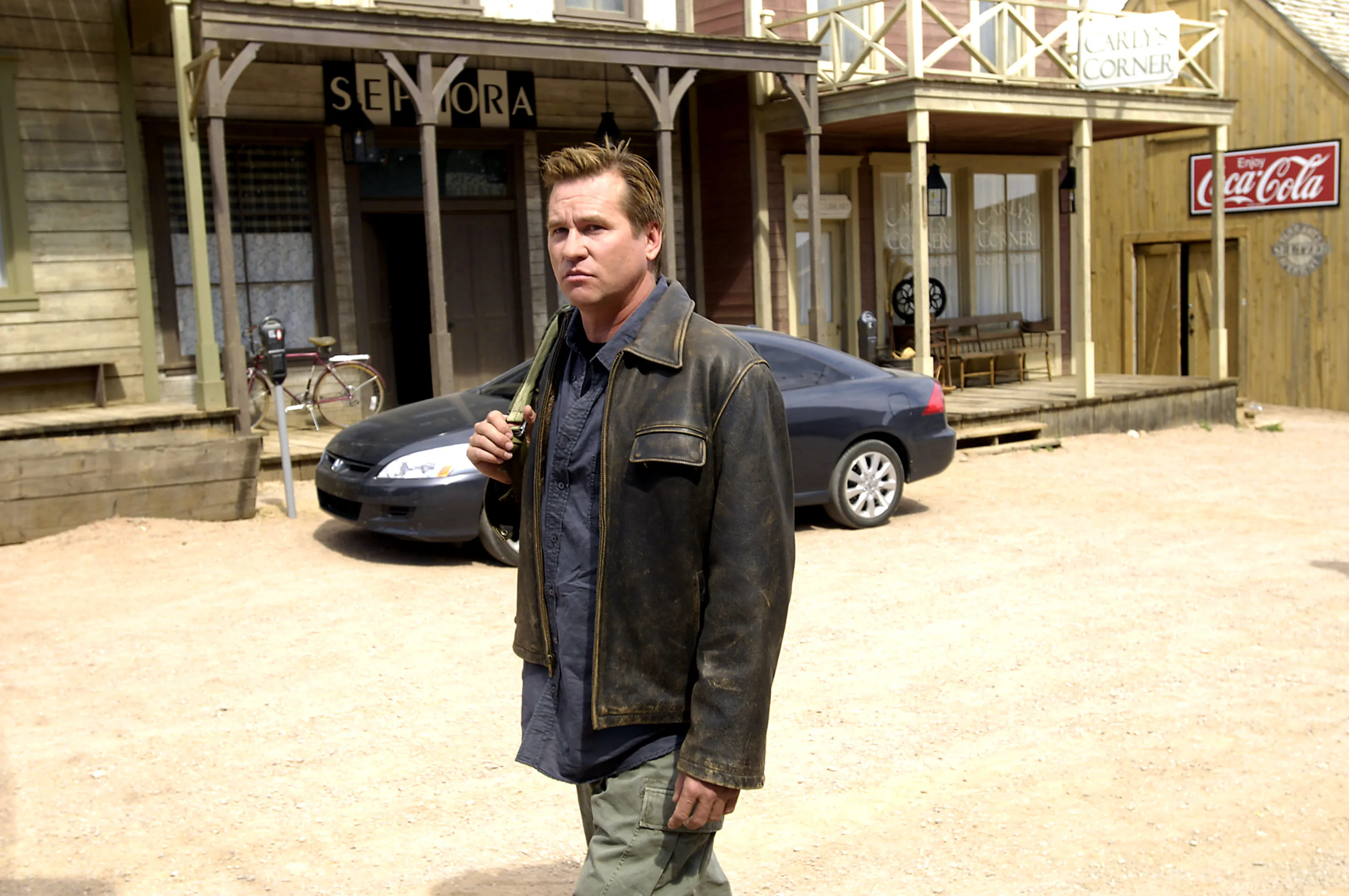 Val Kilmer in a leather jacket stands in a dirt road in front of wooden buildings.