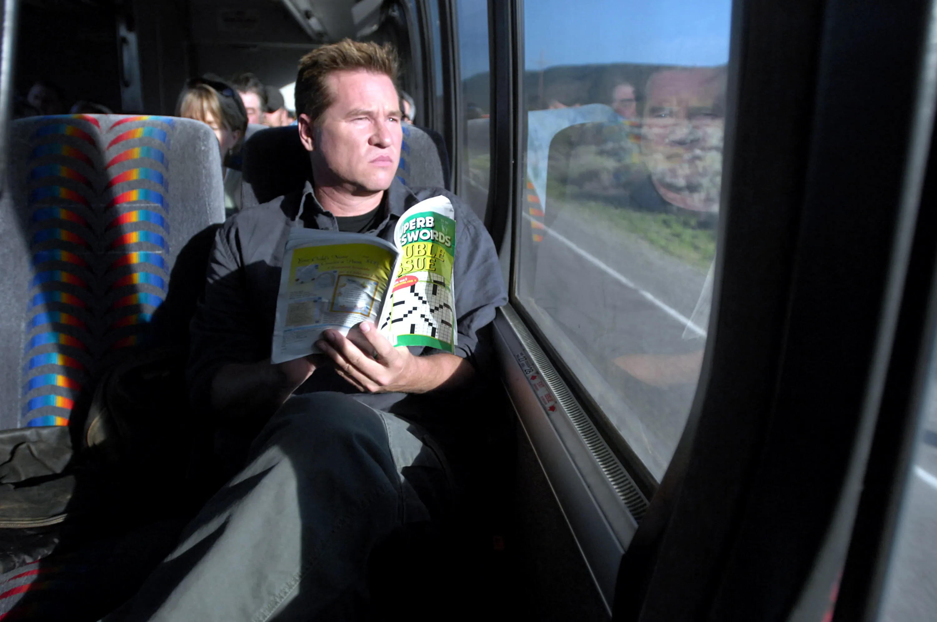 Val Kilmer as a passenger on a bus, holding a crossword puzzle magazine and looking out the window.