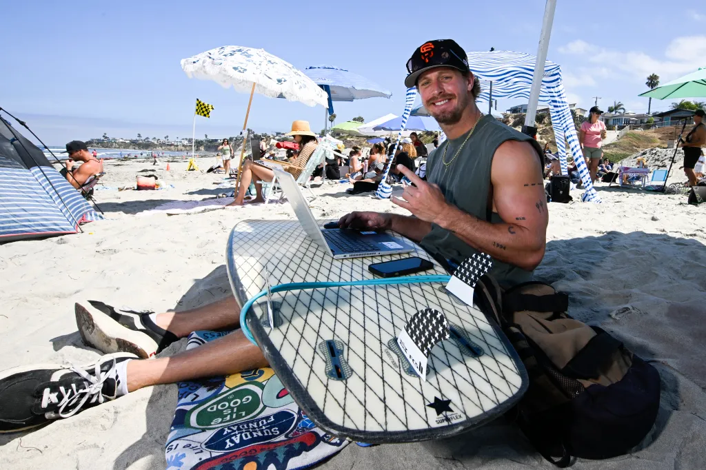 Man working on a laptop on the beach with a surfboard in front of him.