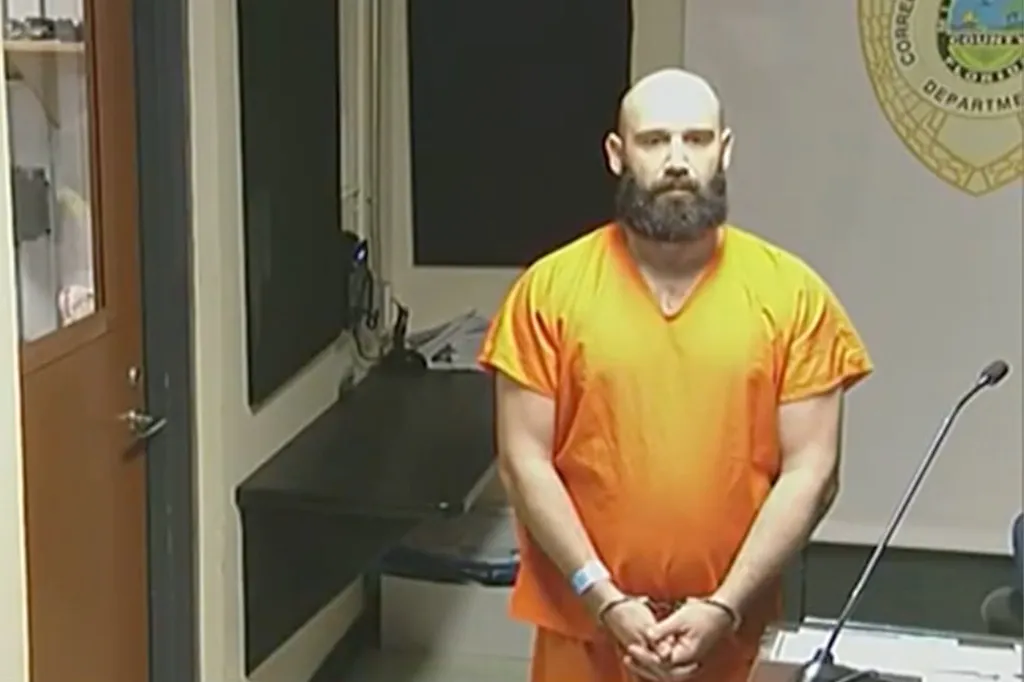 Corey Hutterli standing in an orange jumpsuit with his hands cuffed, with a seal on the wall behind him.