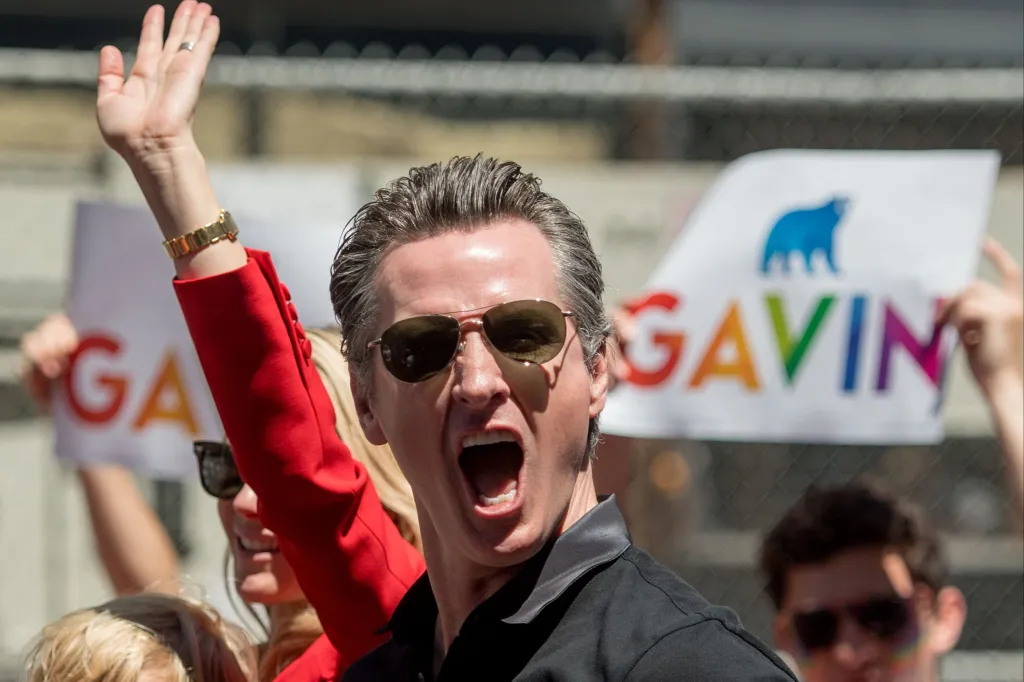 Gavin Newsom reacts with his mouth open while wearing sunglasses and a black collared shirt, with a hand raising on the left and a