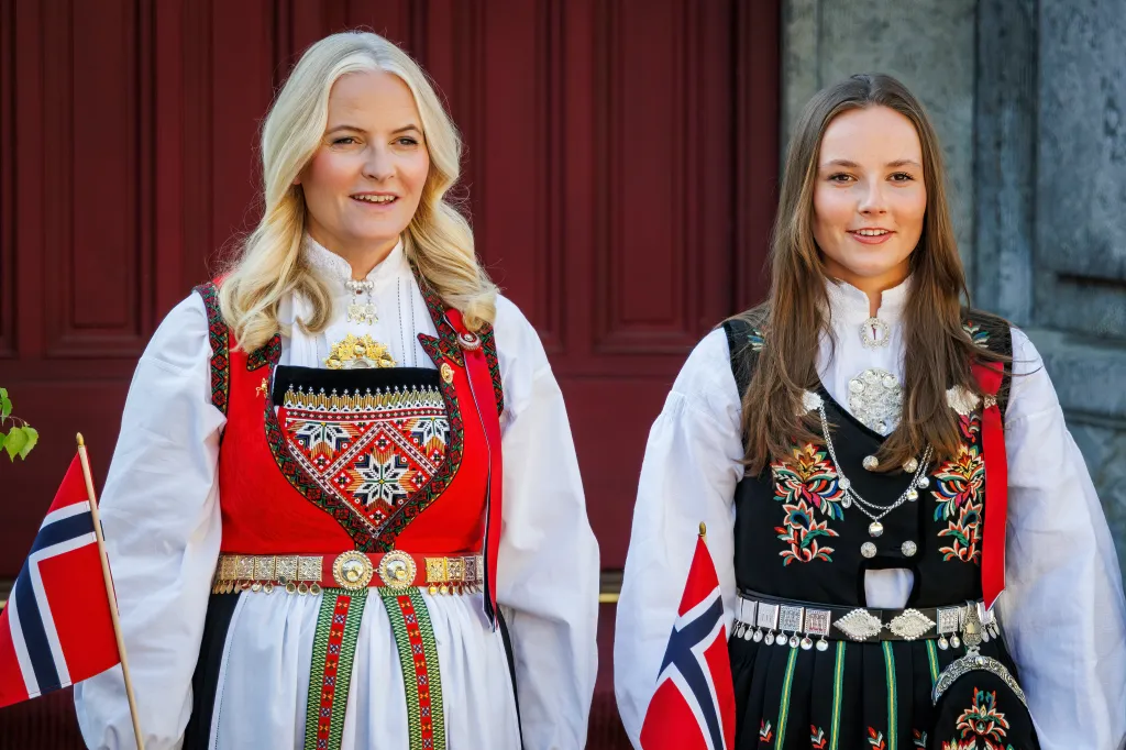 Crown Princess Mette-Marit and Princess Ingrid Alexandra of Norway wearing traditional Norwegian bunad attire and holding Norwegian flags.