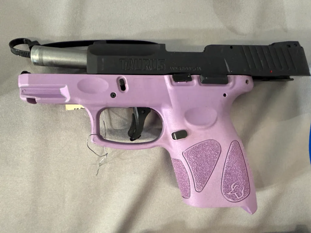 A pink Taurus brand handgun with a black slide, secured with a black zip tie through its barrel, lying on a light-colored surface.