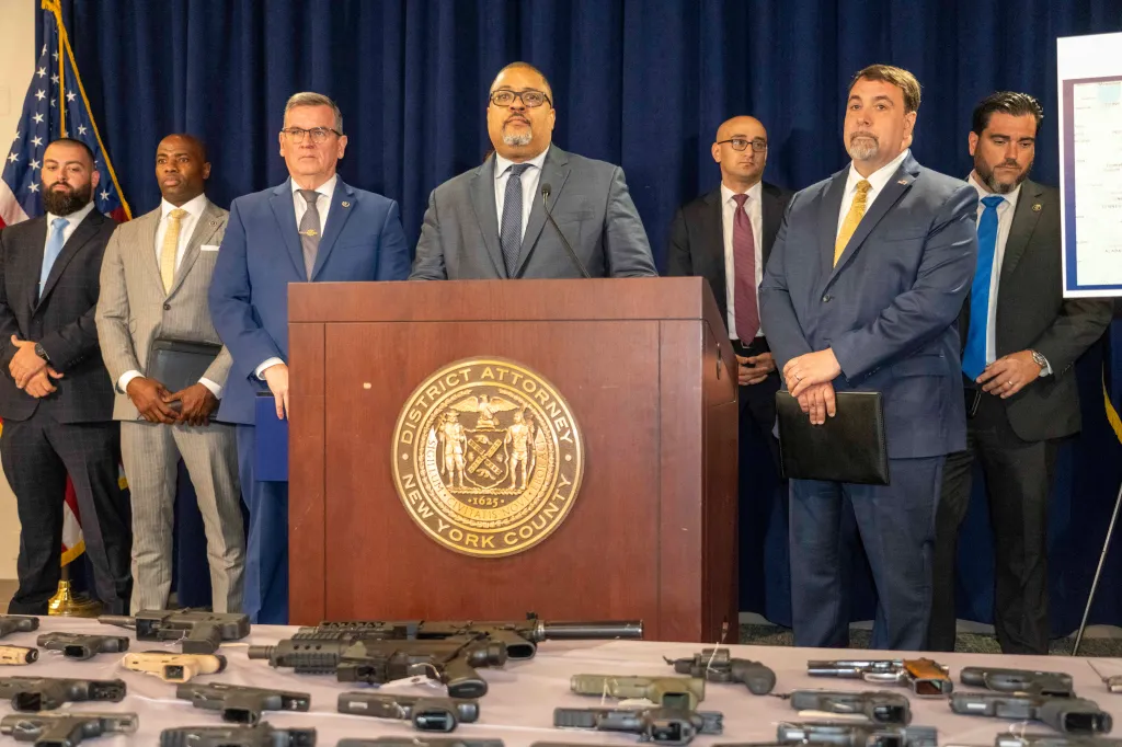 District Attorney Alvin Bragg speaking at a press conference with other officials as guns seized from a trafficking operation are displayed on a table in front of them.