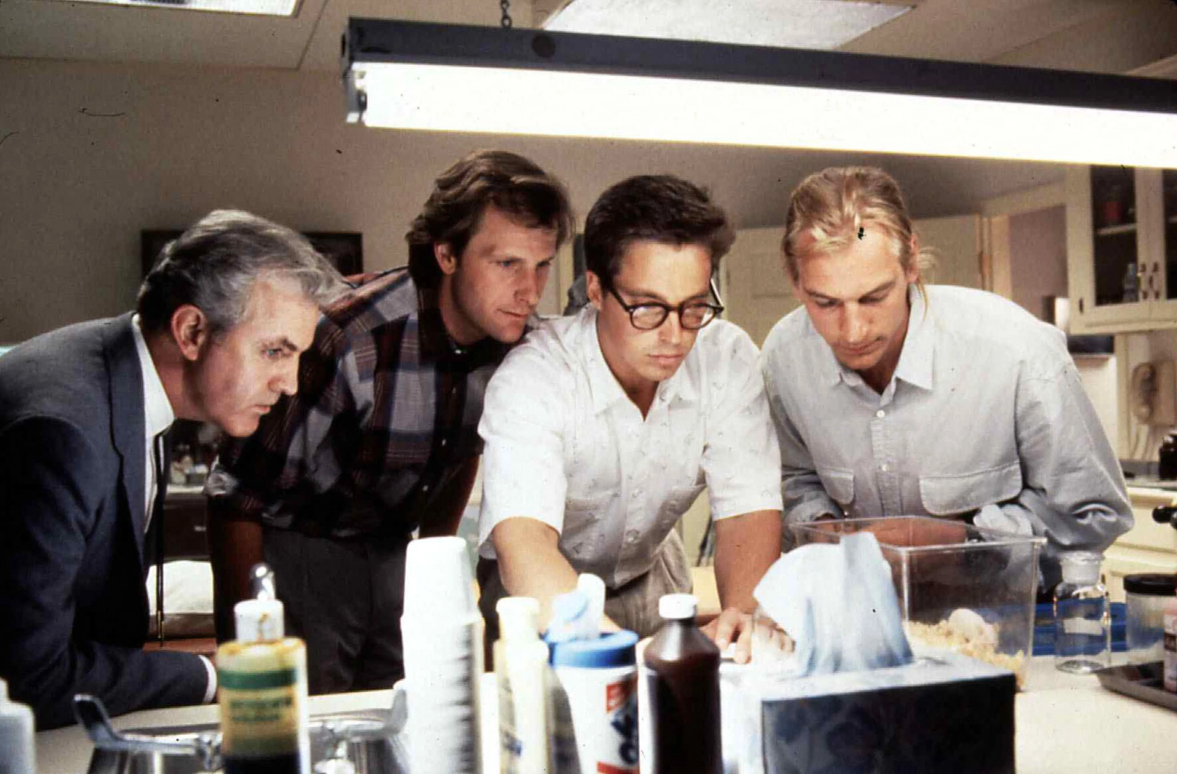 James Handy, Jeff Daniels, Brian McNamara, and Julian Sands leaning over a laboratory counter in a still from the film 
