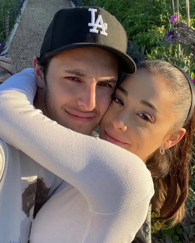 Dalton Gomez, in a black LA baseball cap, and Ariana Grande, in a white top, hugging and smiling at the camera.