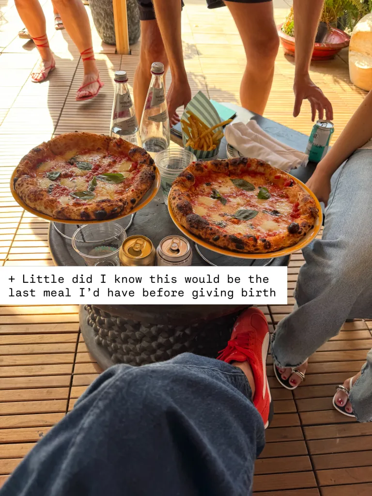 A high-angle shot of two large round pizzas, fries, and drinks on a table, surrounded by people's legs and feet.