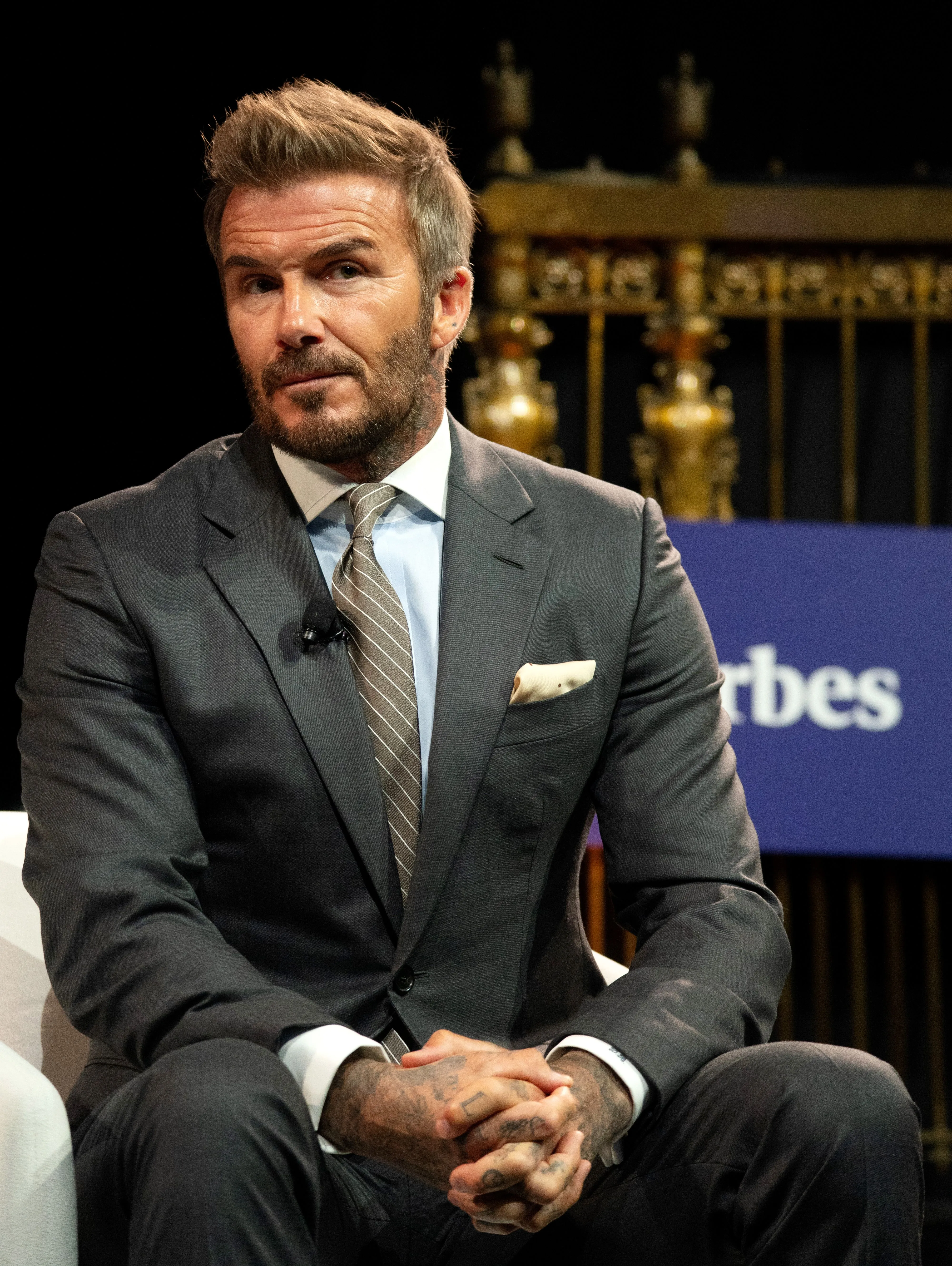 David Beckham in a gray suit and striped tie, seated with hands clasped, addressing an audience.