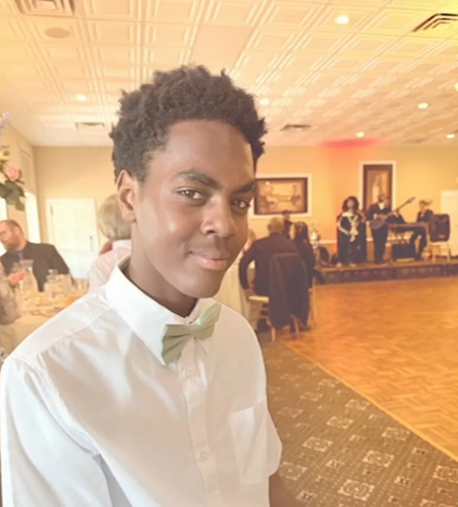 A young Black man wearing a white shirt and light green bow tie smiles at the camera, with a band performing in the background.