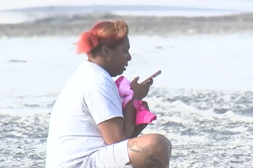 Davoris Carter's mother clutching swim trunks she found on the shoreline, holding a phone while sitting at the beach.