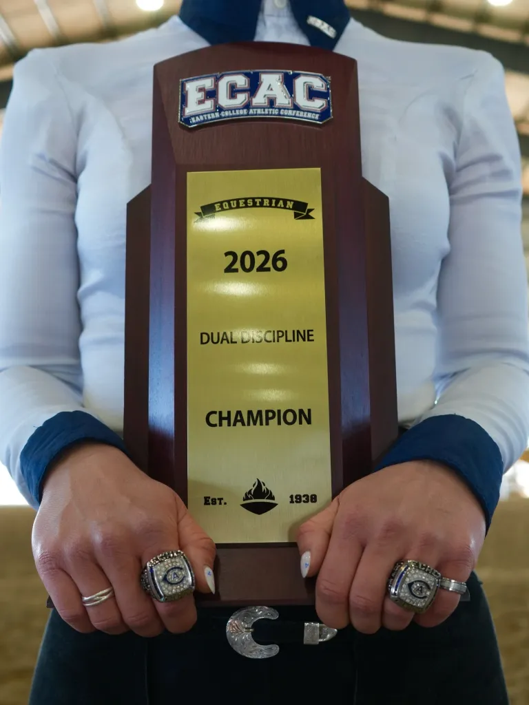 A person holding an ECAC Equestrian 2026 Dual Discipline Champion trophy, with two championship rings on their fingers.