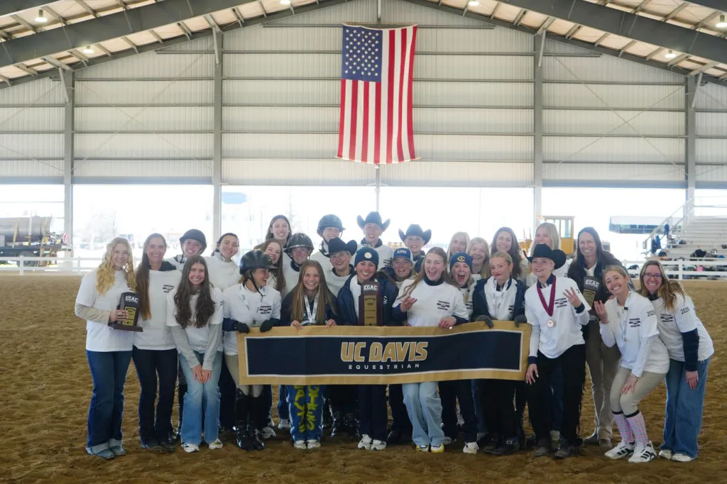 The UC Davis Equestrian team celebrates their conference championship with a banner and trophy, standing under an American flag in an arena.