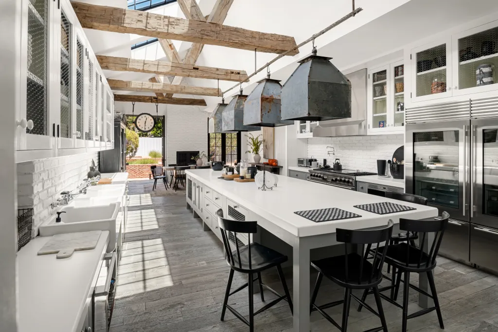 Large open-plan kitchen with white cabinetry, a long central island with seating, exposed wooden beams, and a view to an outdoor patio.