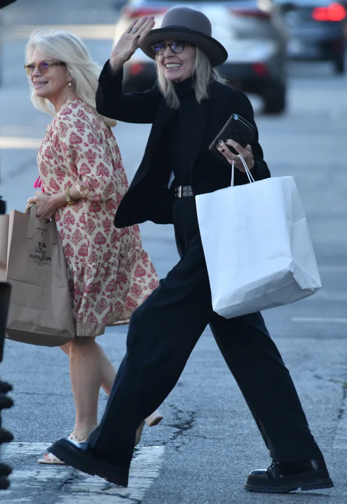 Diane Keaton wearing a hat, glasses, and a black outfit, waving as she walks with a friend.