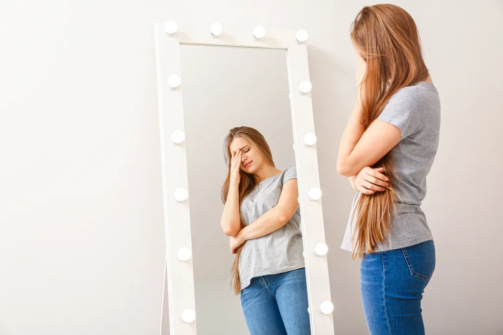 A displeased young woman looks in a mirror.