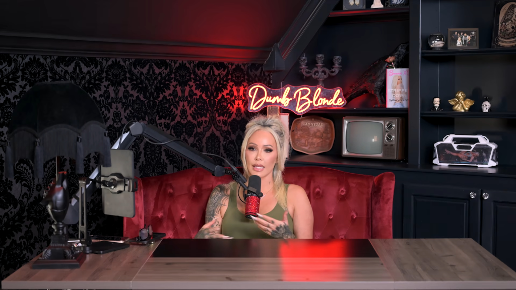 Bunnie XO speaking into a microphone while seated on a red velvet couch in a room with a 