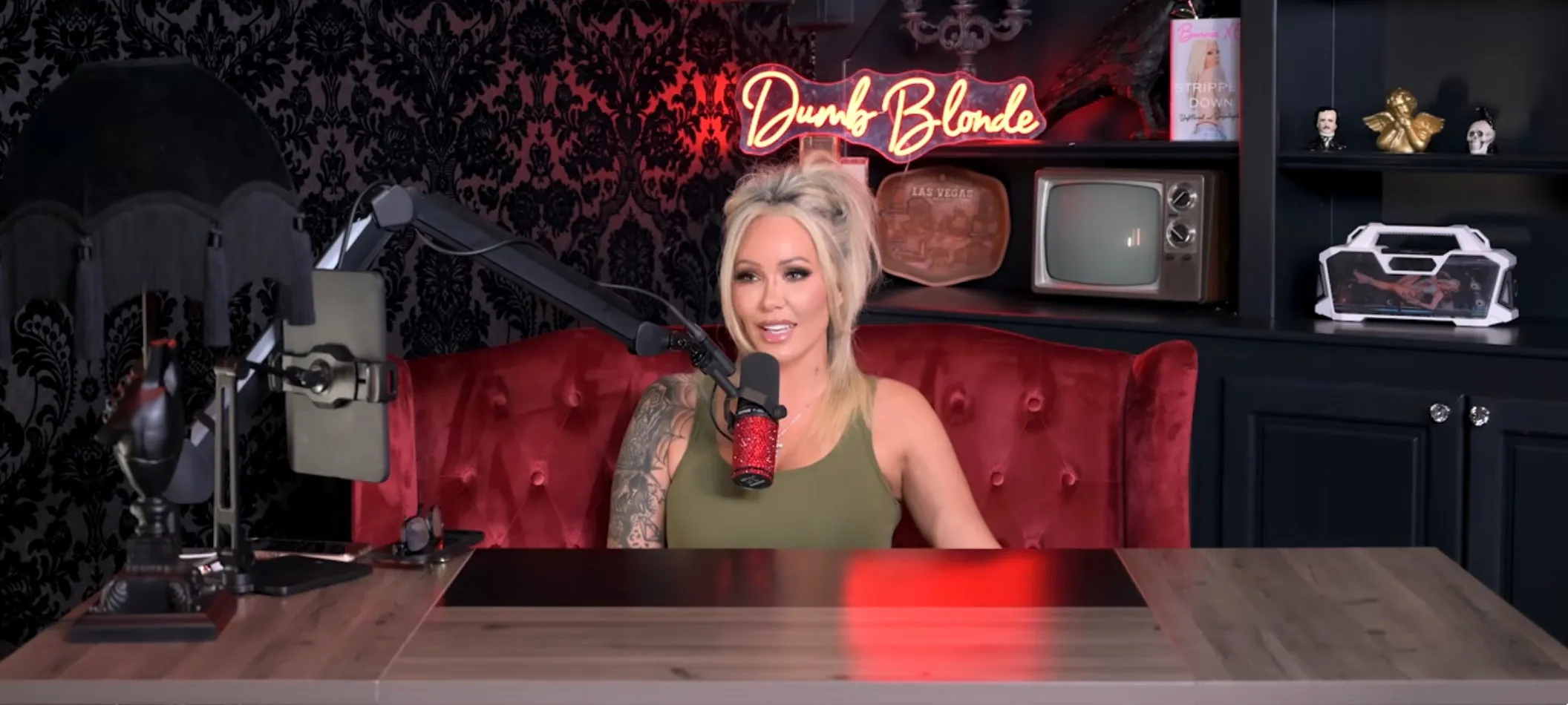 A woman with blonde hair and tattoos sits in a red chair in front of a microphone, with a