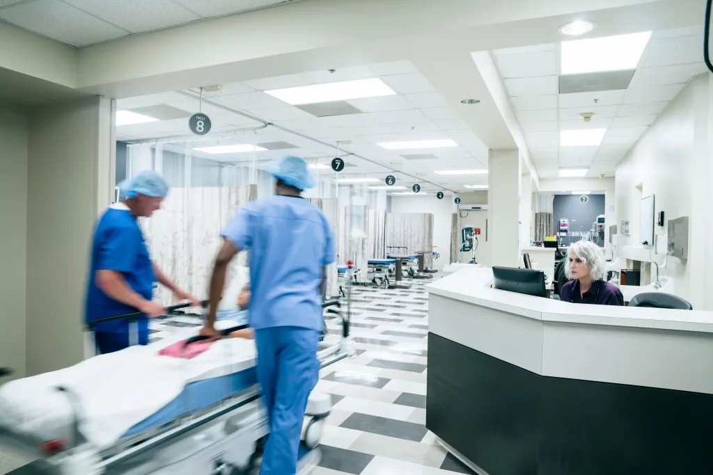 Two doctors push a patient on a gurney through a hospital hallway as a woman works at a reception desk.