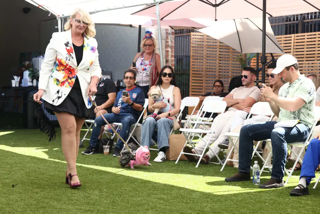 A blonde woman in a floral blazer walks a pink dog down a runway for a dog fashion show.