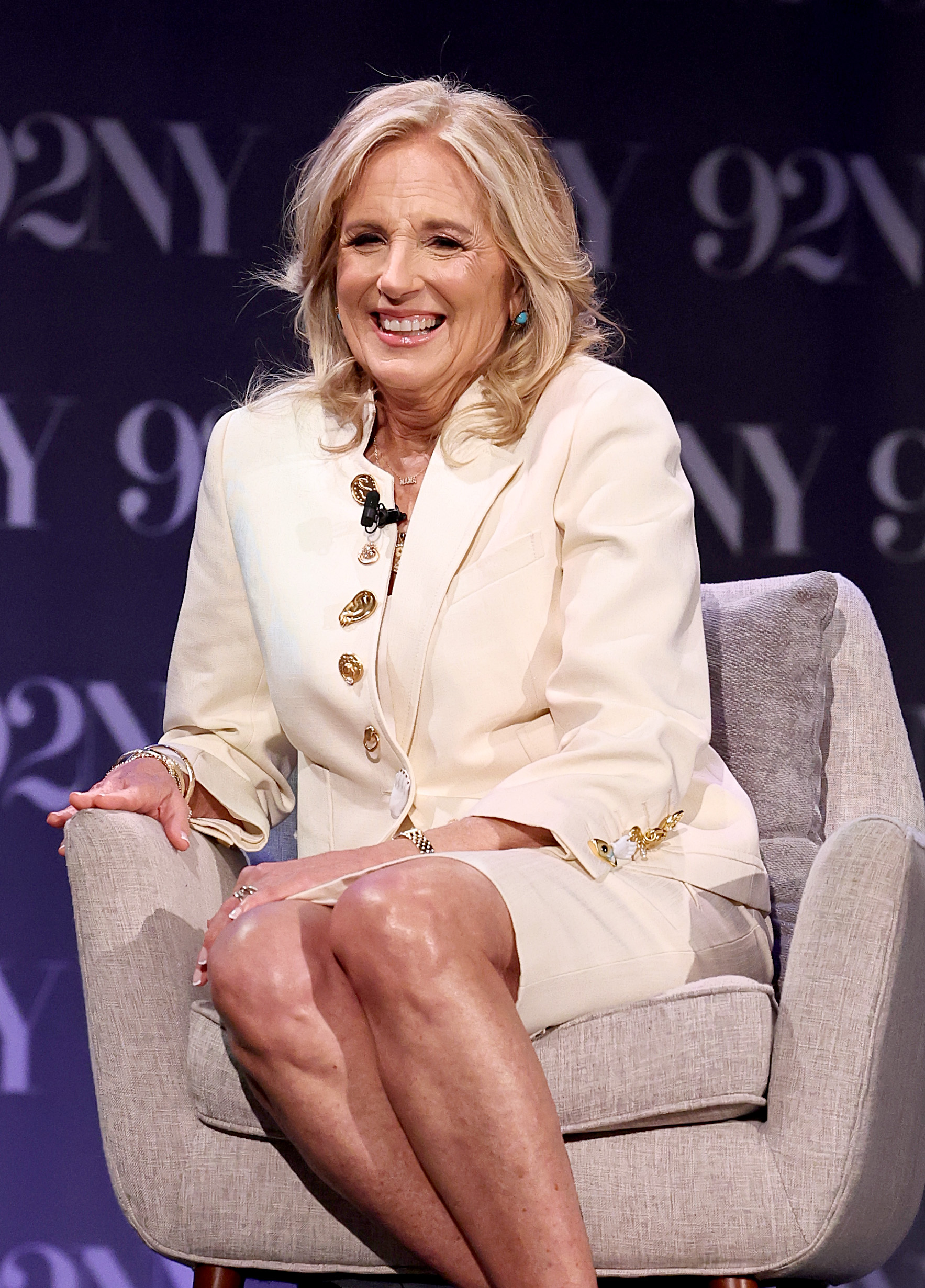 Dr. Jill Biden sits on stage in a cream suit with unique gold buttons, smiling and laughing at an event in New York City.