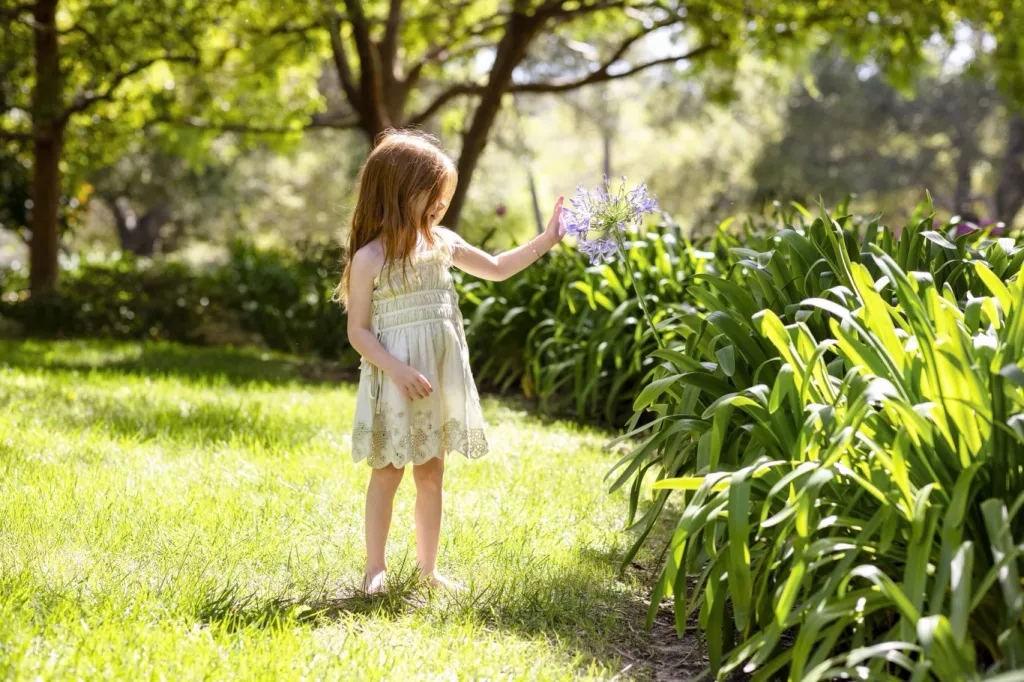 Lilibet, a young girl with red hair, in a light-colored dress and bare feet, touches a purple flower in a garden.