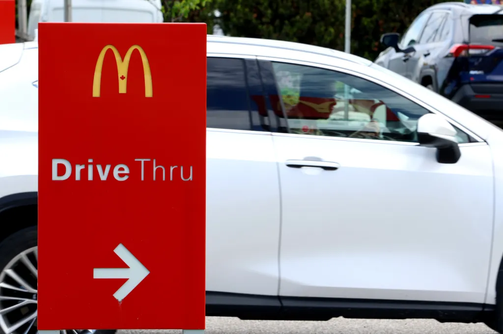 A red McDonald's drive-thru sign with a maple leaf on the golden arches, with a white car in the background.