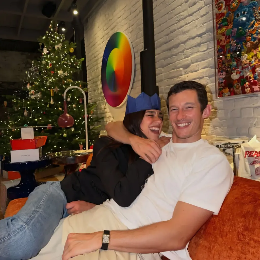 Dua Lipa and Callum Turner embracing and laughing, with a Christmas tree in the background.