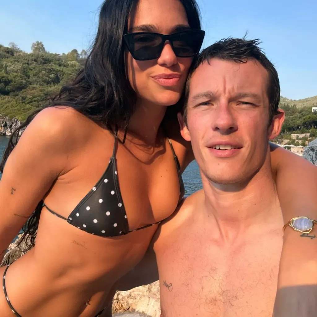 Dua Lipa and Callum Turner embracing in a sunny outdoor setting.