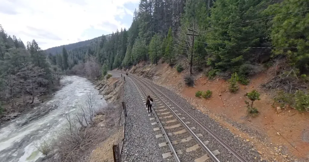 A person walks along a train track next to a flowing river, surrounded by forested hills.